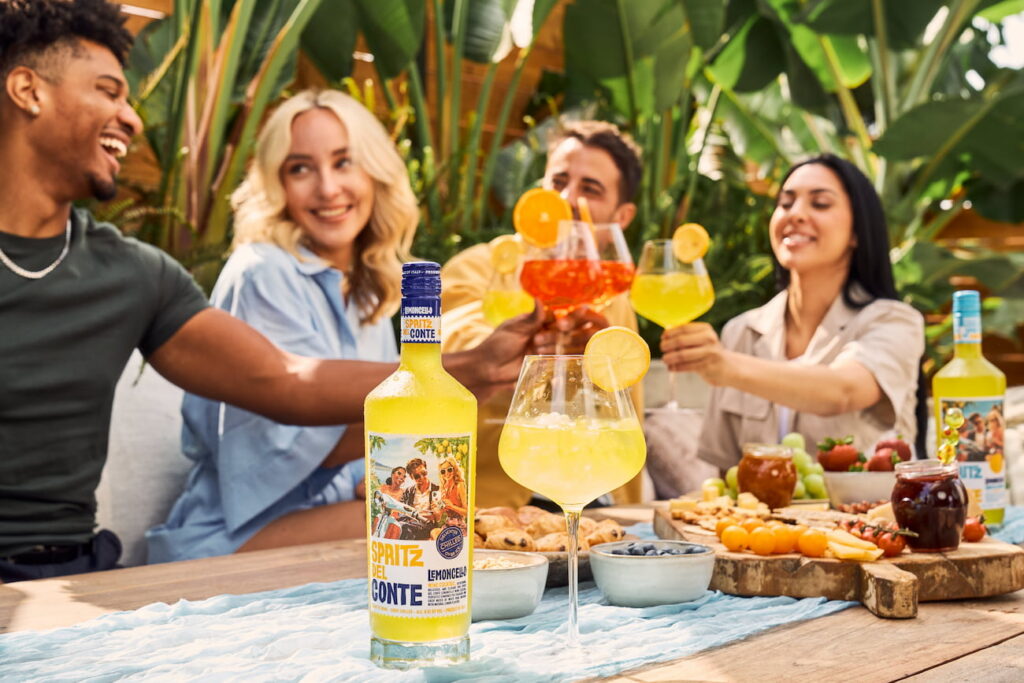 Friends toast with Spritz Del Conte Lemoncello Spritz at a summer gathering featuring oranges, berries, grapes, cheeses, crackers, pastries, preserves, and other light bites.